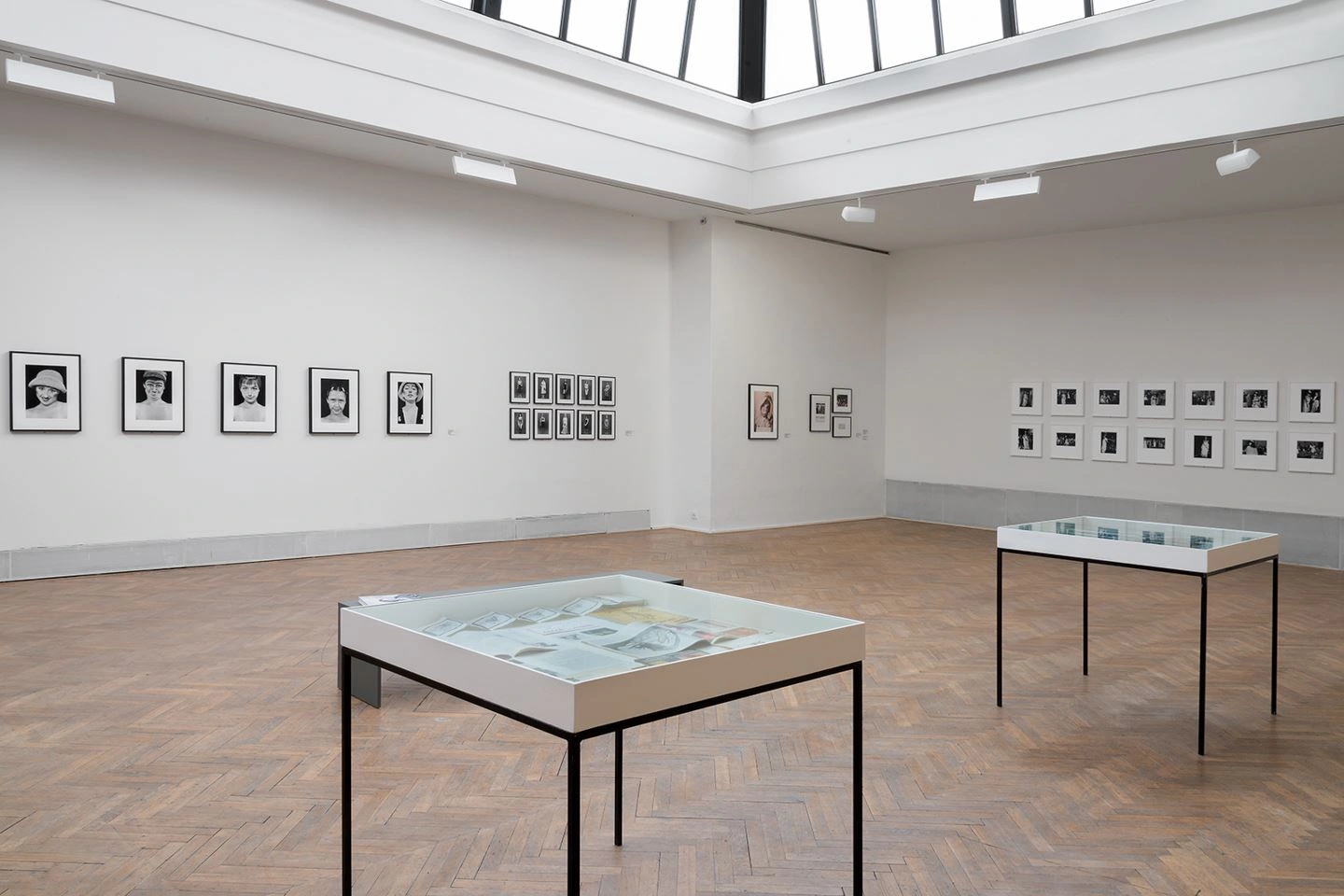 A bright exhibition room with photographs on the wall and a parquet floor can be seen. In the room are two display cases with black table legs