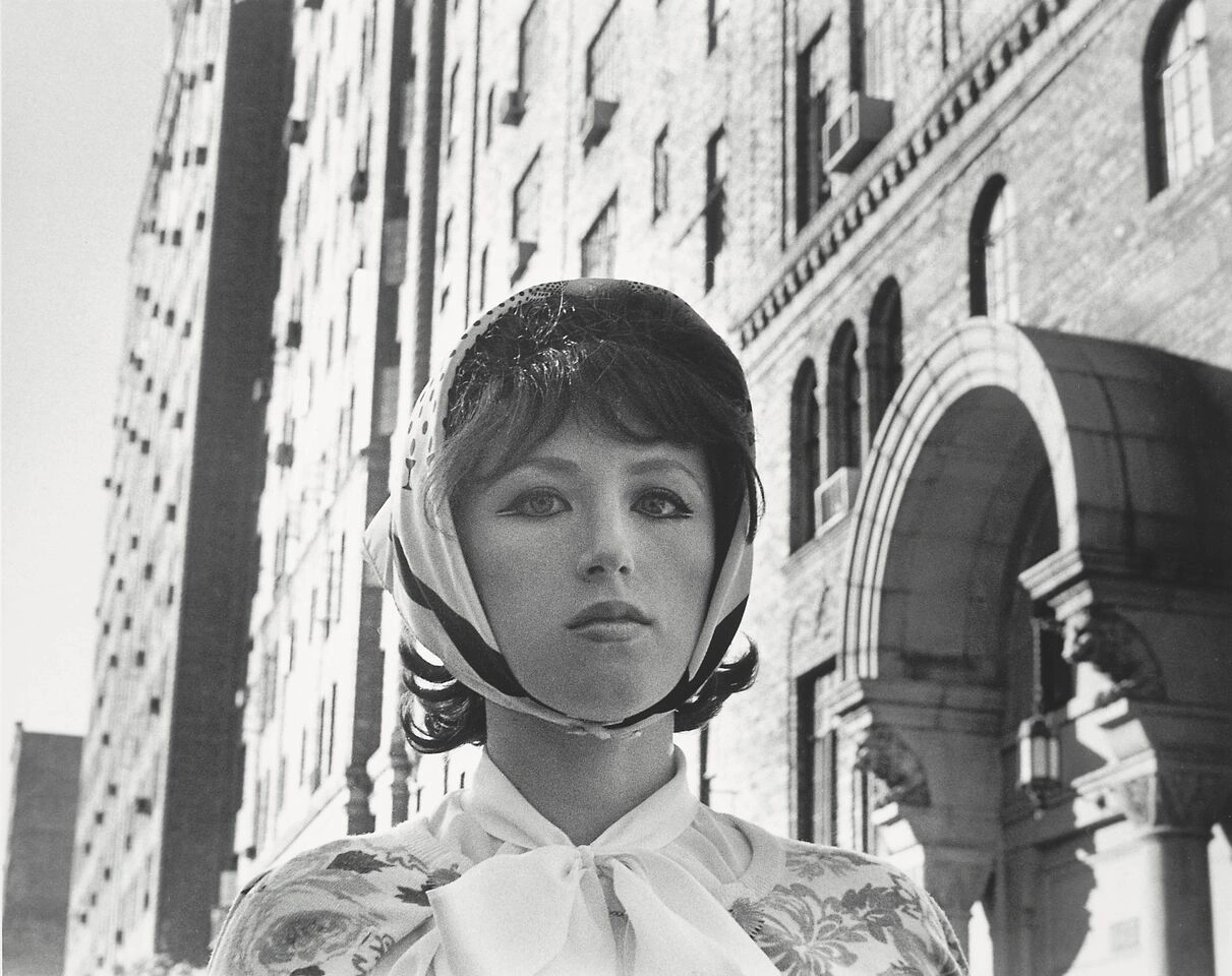 Das Selbstportrait von Cindy Sherman aus dem Jahr 1978 zeigt die Künstlerin in schwarz /weiß. Man sieht nur ihren Oberkörper. Sie trägt einen auffälligen Kajalstrich, eine Perücke mit Kopftuch und eine Schluppenbluse. Im Hintergrund sieht herrschaftlich anmutende Häuser. Sie blick über die Kamera hinweg.