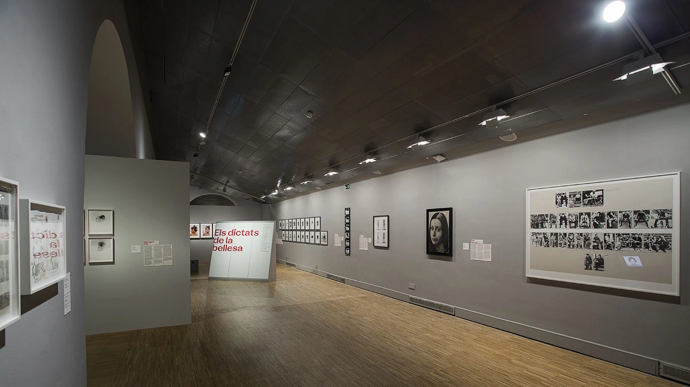 View inside an art gallery with an arched ceiling and parquet flooring, with framed black-and-white photos hanging on the walls, and an open doorway in the middle leading to another exhibition room with the inscription "Els silencis clamorosos".