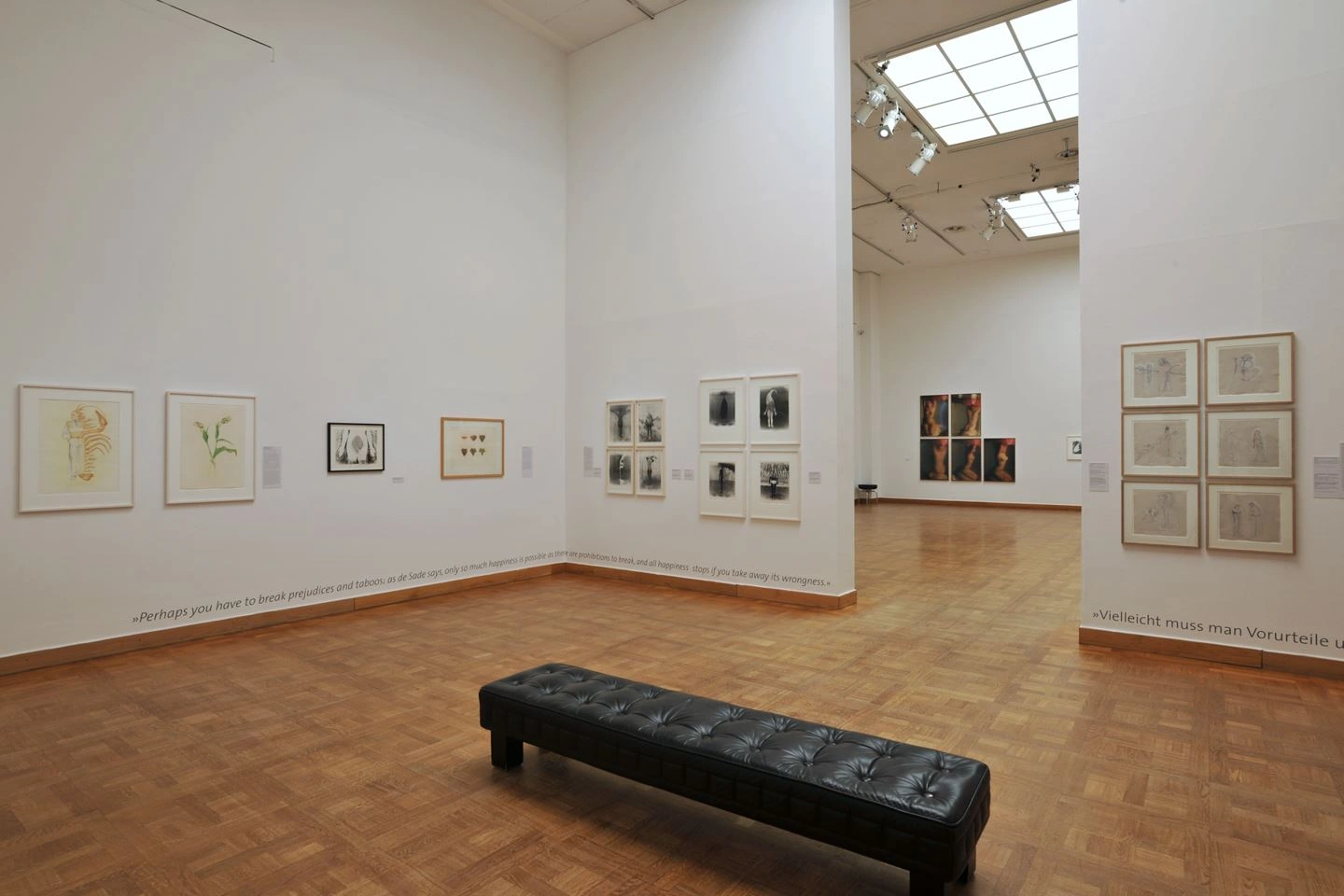 Empty exhibition space featuring a collection of artworks on white walls with a black bench in the center on a wooden parquet floor, daylight streaming through the skylight.