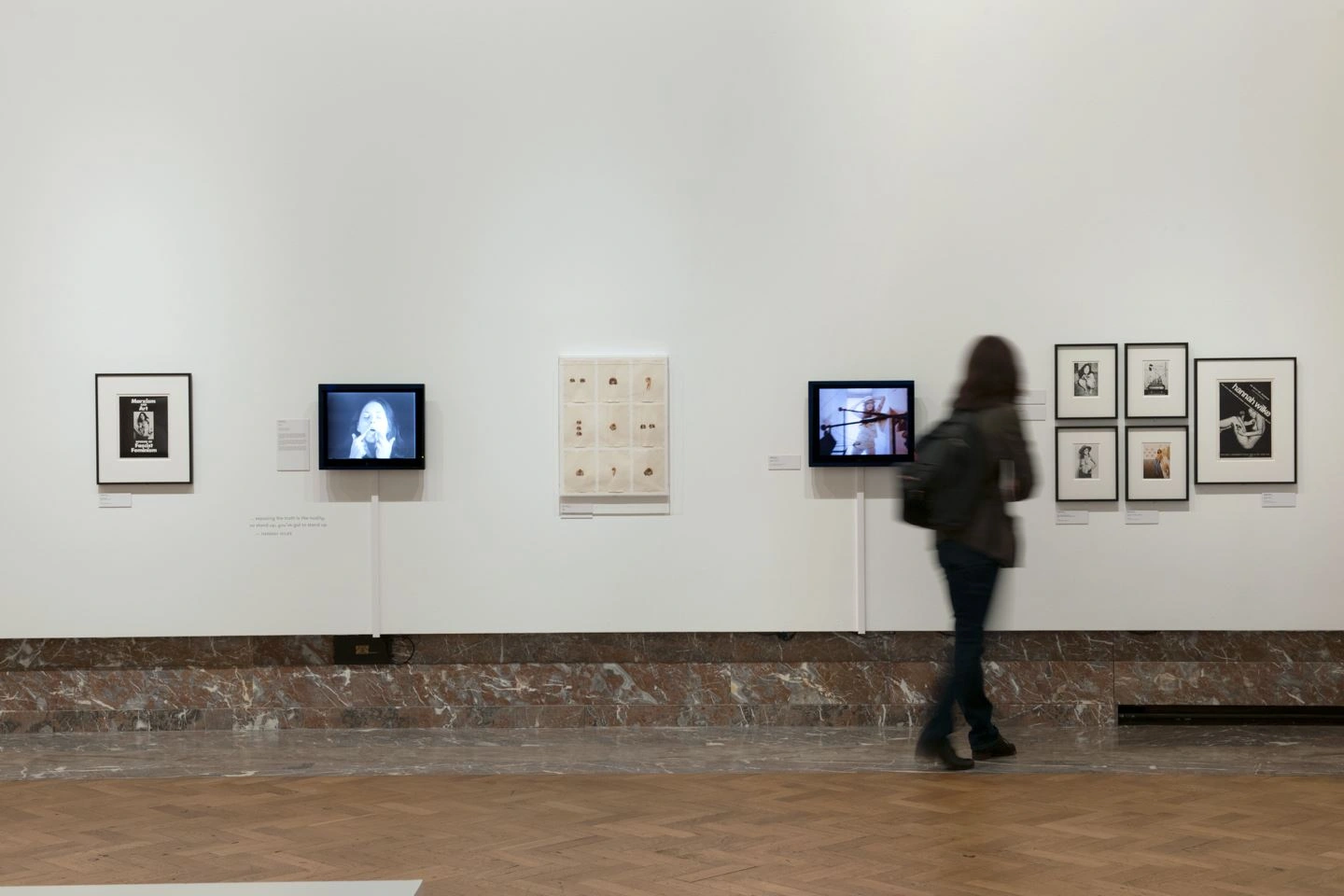 Several works of art by Bozar are hung on a white wall in an exhibition hall. A visitor looks at the pictures and can be seen from behind.