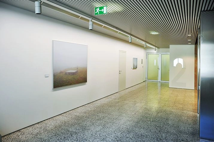 Contemporary art gallery with a minimalist design, featuring two large paintings on the left wall, a granite-colored floor, and a modern suspended ceiling.