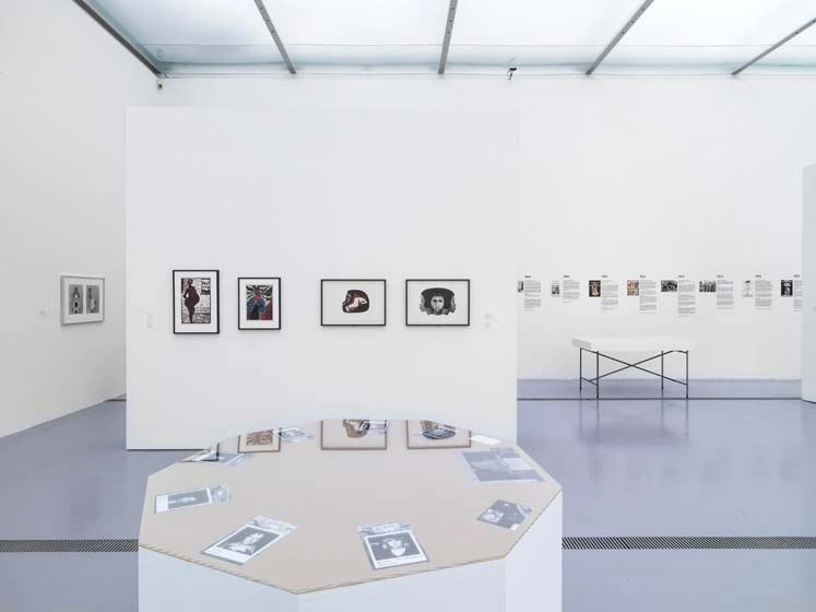 A very modern and bright exhibition room shows photographs in black frames on the walls. There are two display tables in the room. One of them mirrors the pictures on the wall behind it