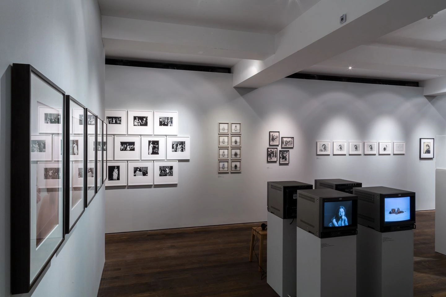 Interior view of a photo art gallery featuring numerous framed black and white photographs on bright walls and multimedia presentation pedestals with screens in an exhibition hall with wooden flooring and modern lighting.