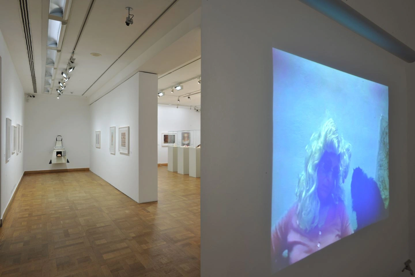 Blick in eine helle Galerie mit holzgetäfeltem Boden, an den Wänden hängen verschiedene gerahmte Bilder. Am Ende des Raumes wird ein großes Videokunstwerk auf einer Wand projiziert, das eine undeutliche Figur unter Wasser zeigt.