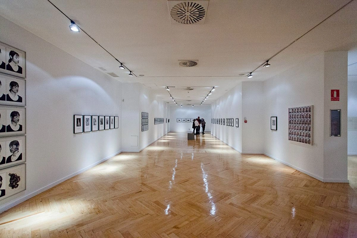 A long exhibition room with exhibits on the walls to the left and right can be seen. Far in the back, visitors are gathered in front of one of the works of art.