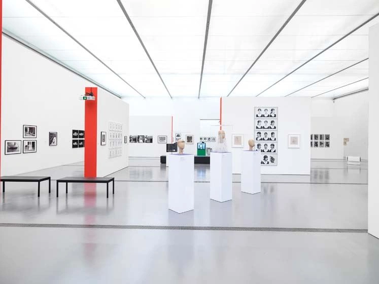 A bright exhibition room with red accentuated wall sides. Photographs hang on the white walls and there are showcases on white pedestals in the room.