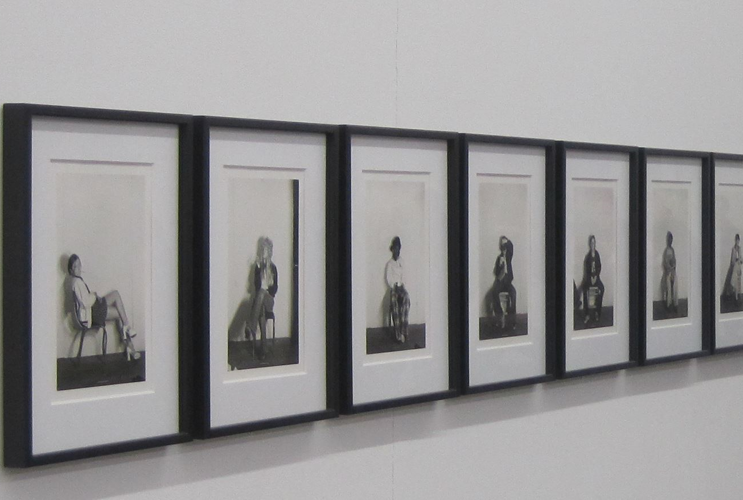 Sequence of framed black-and-white photographs on a white wall in an art gallery, depicting a series of portraits.