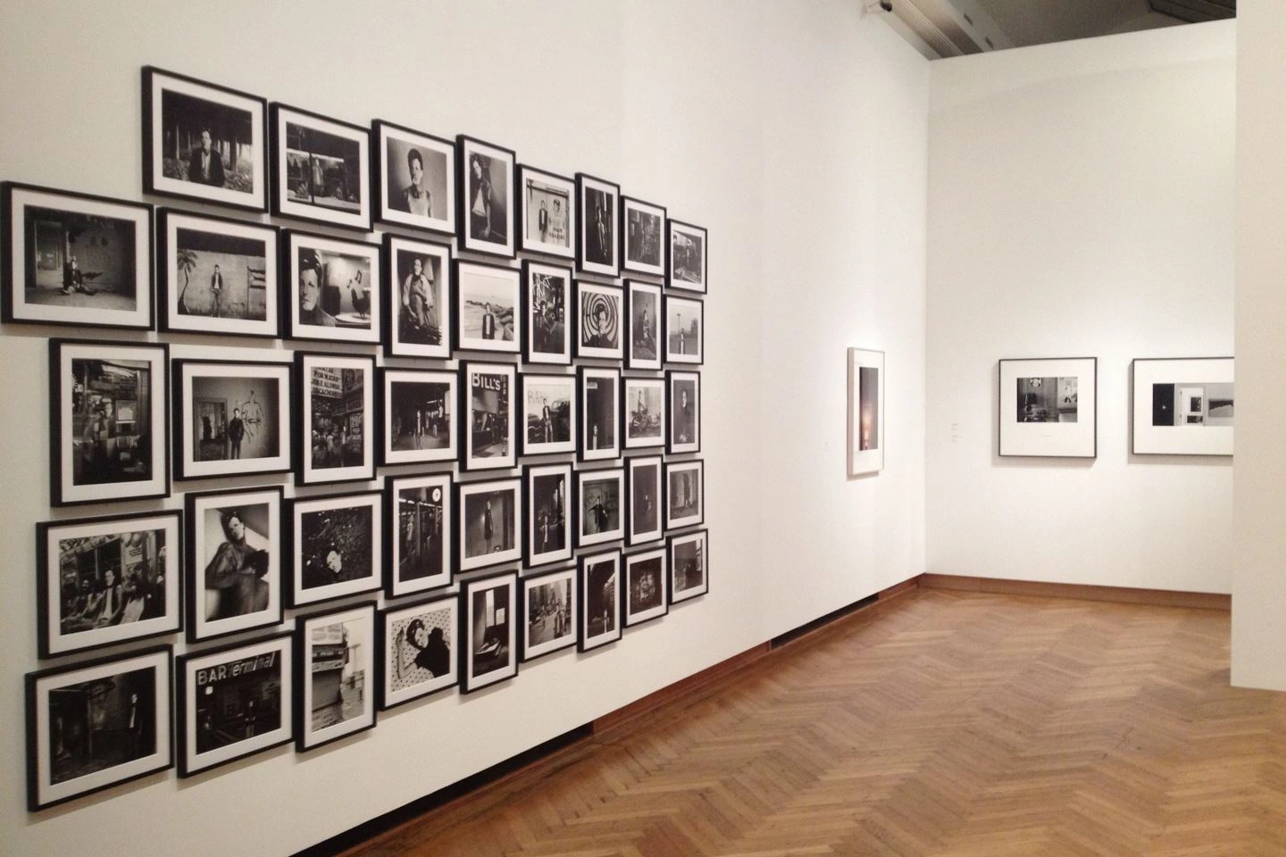 Gallery corridor featuring a montage of framed black-and-white photographs on a white wall extending around the corner, set on parquet flooring.