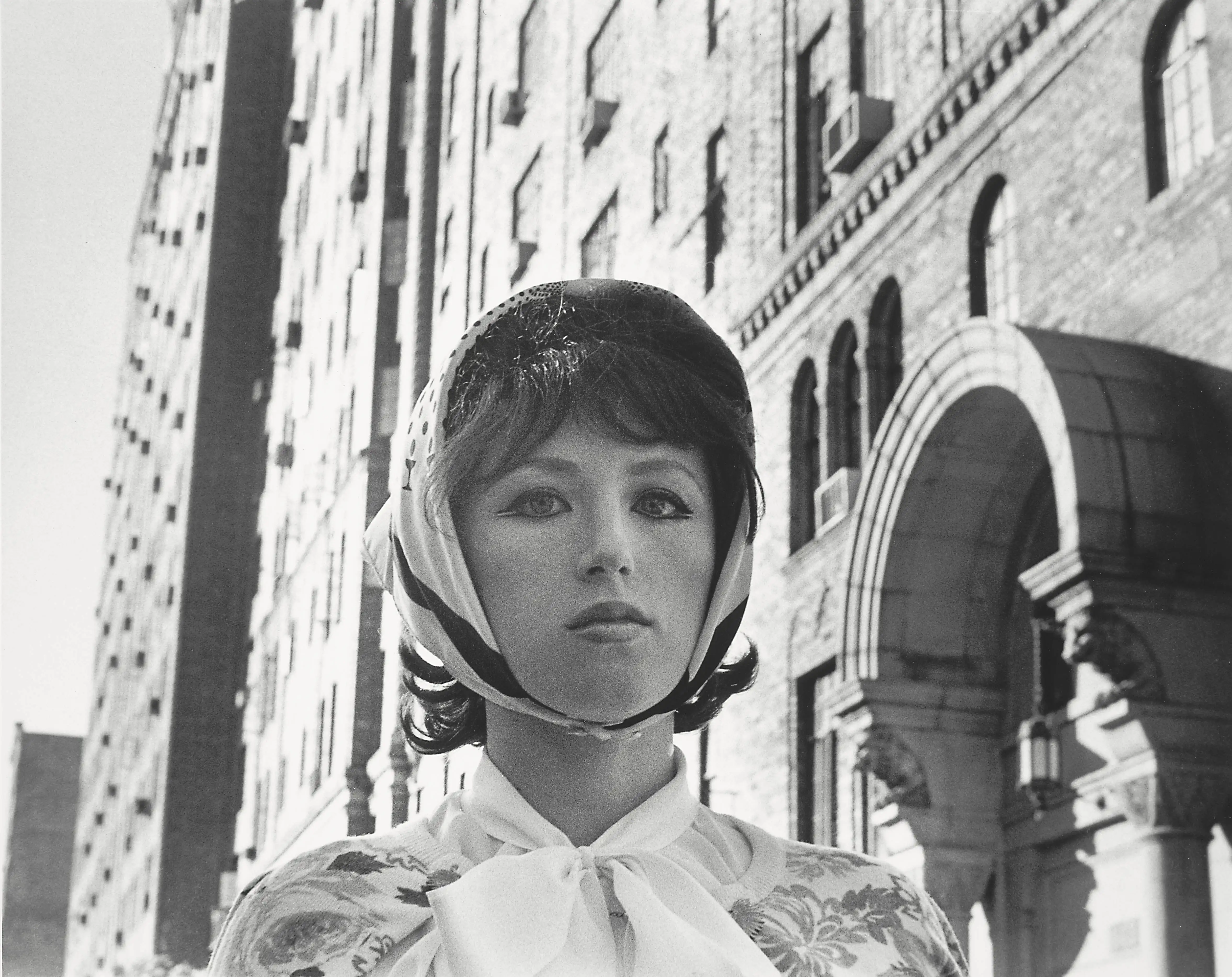 This self-portrait by Cindy Sherman from 1978 shows the artist in black and white. Only her upper body is visible. She is wearing eye-catching eyeliner, a wig with a headscarf and a blouse. In the background are stately-looking houses. She looks over the camera.