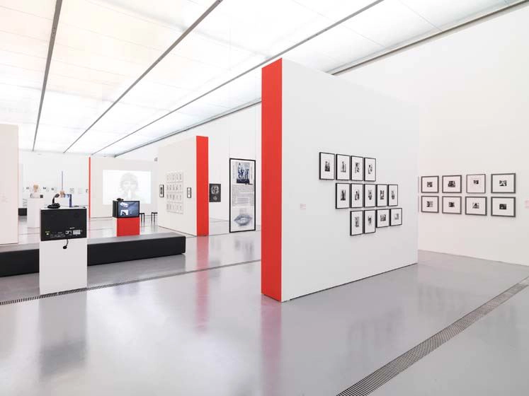 In a bright exhibition room there are several partition walls, the side wall of which is painted in bright red. Black and white photographs hang on all the walls. Further back in the room are monitors and a black seat without backrests.