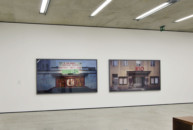 Bright gallery with two large cityscape paintings on a white wall and a wooden bench in the foreground.