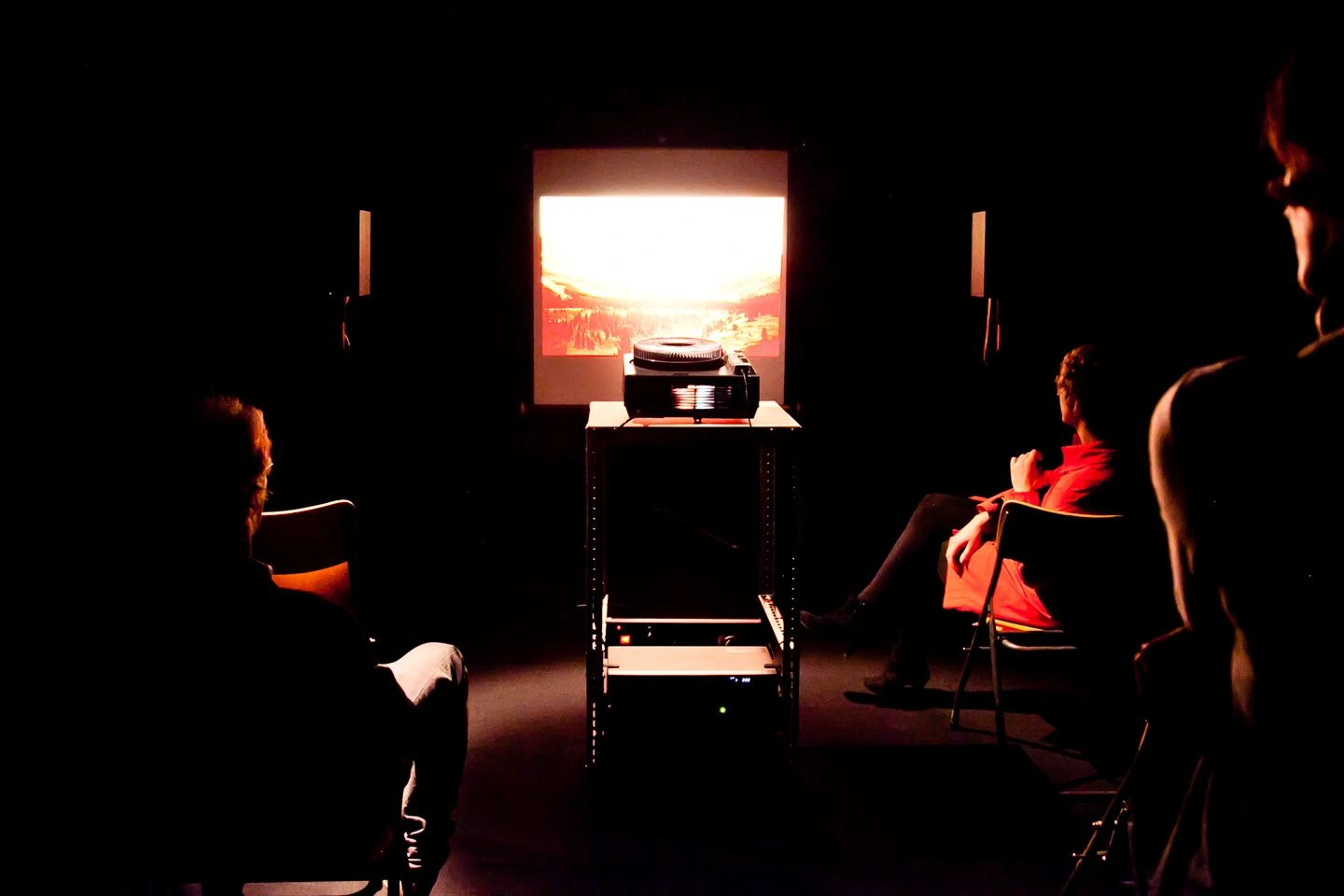 Audience seated in a dimly lit room watching a slide projection of a sunset on a screen.