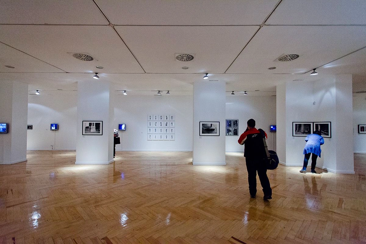 Eine Totalaufnahme eines großen Ausstellungsraumes mit weißen Wänden und Holzfussboden ist zu sehen. Im Raum stehen mehrere Besucher und betrachten unterschiedliche Bilder.