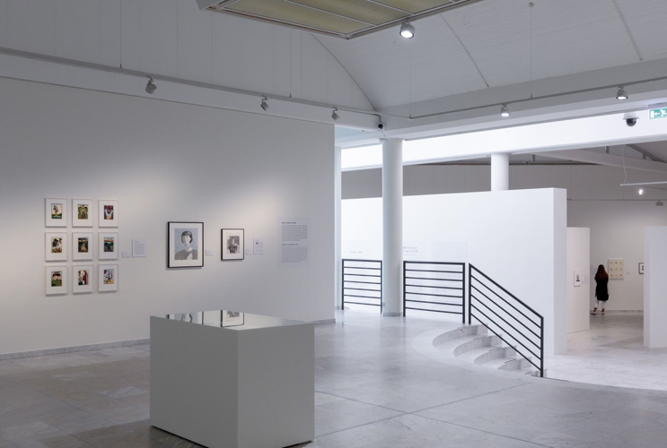 A close-up of a very bright exhibition room can be seen. To the right of it is a stair railing. The exhibition space is divided several times by partition walls. Works of art are hung on the partition walls. Lamps with spotlights are attached to the ceiling of the room.