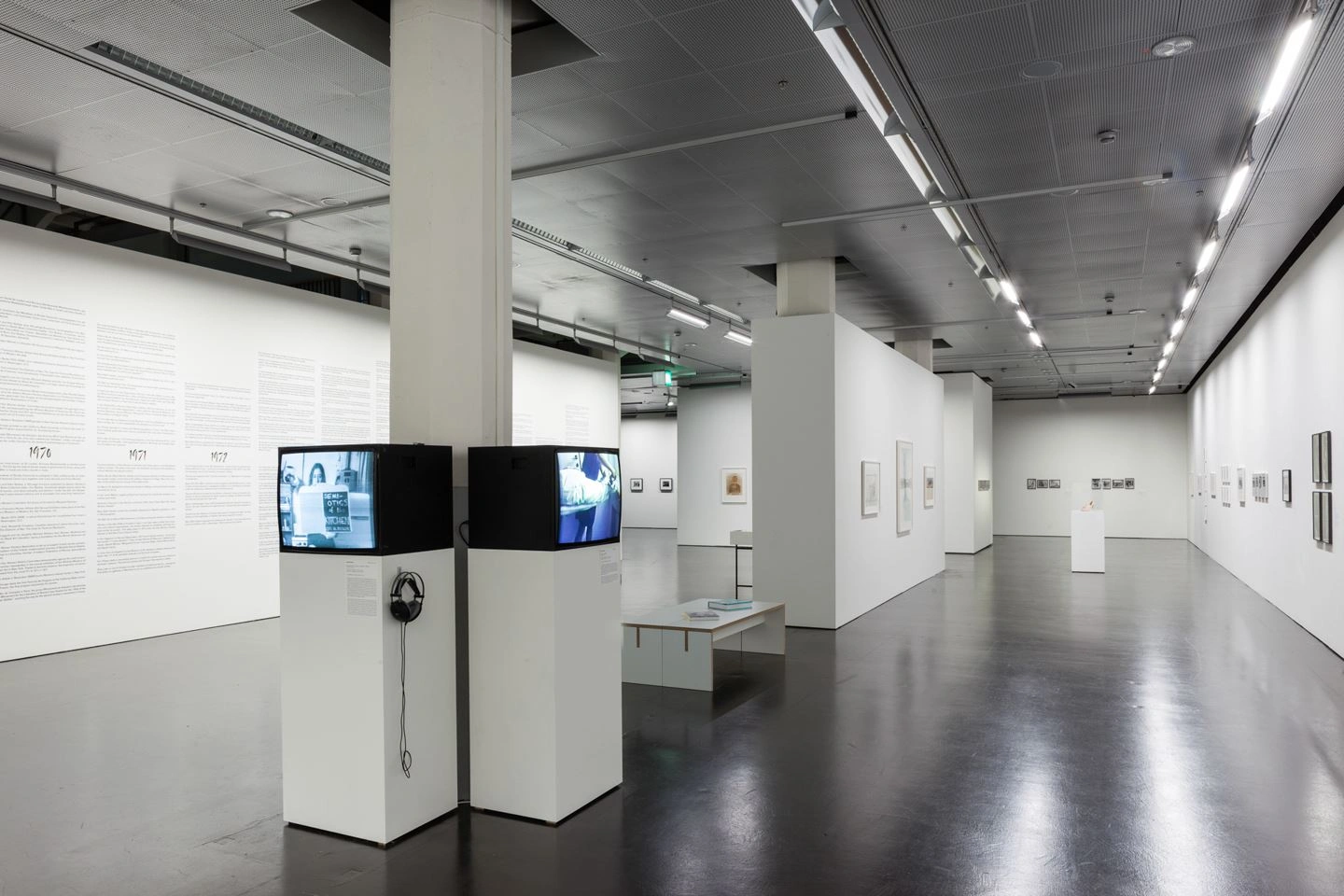 Ein durch eine Trennwand geteilter großer Ausstellungsraum ist zu sehen. An allen Wänden sind Kunstwerke abgebildet. Im vorderen Bereich des Raumes in der Mitte sind zwei Sockel mit Monitoren aufgestellt, die unterschiedliche Videos abspielen.