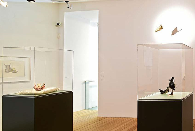 Panoramic view of a brightly-lit art gallery with parquet flooring, featuring three display cases with artistic objects, and a framed wall drawing on the right, next to a staircase leading to an upper floor.