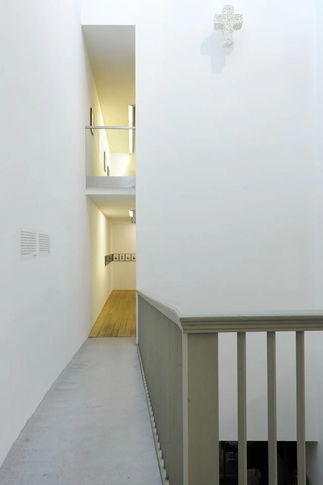 Bright corridor leading to an exhibition room with several portrait-format paintings.