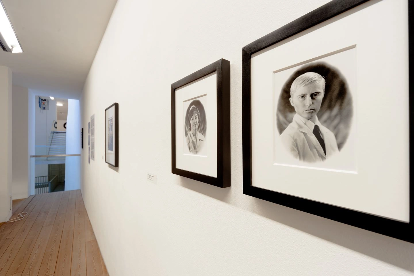 Auf einer weißen Wand hängen zwei Portraitfotos, rechts ein junger Mann mit hellen Haaren und weißem Jacket, links eine junge Dame mit dünkleren Haaren.