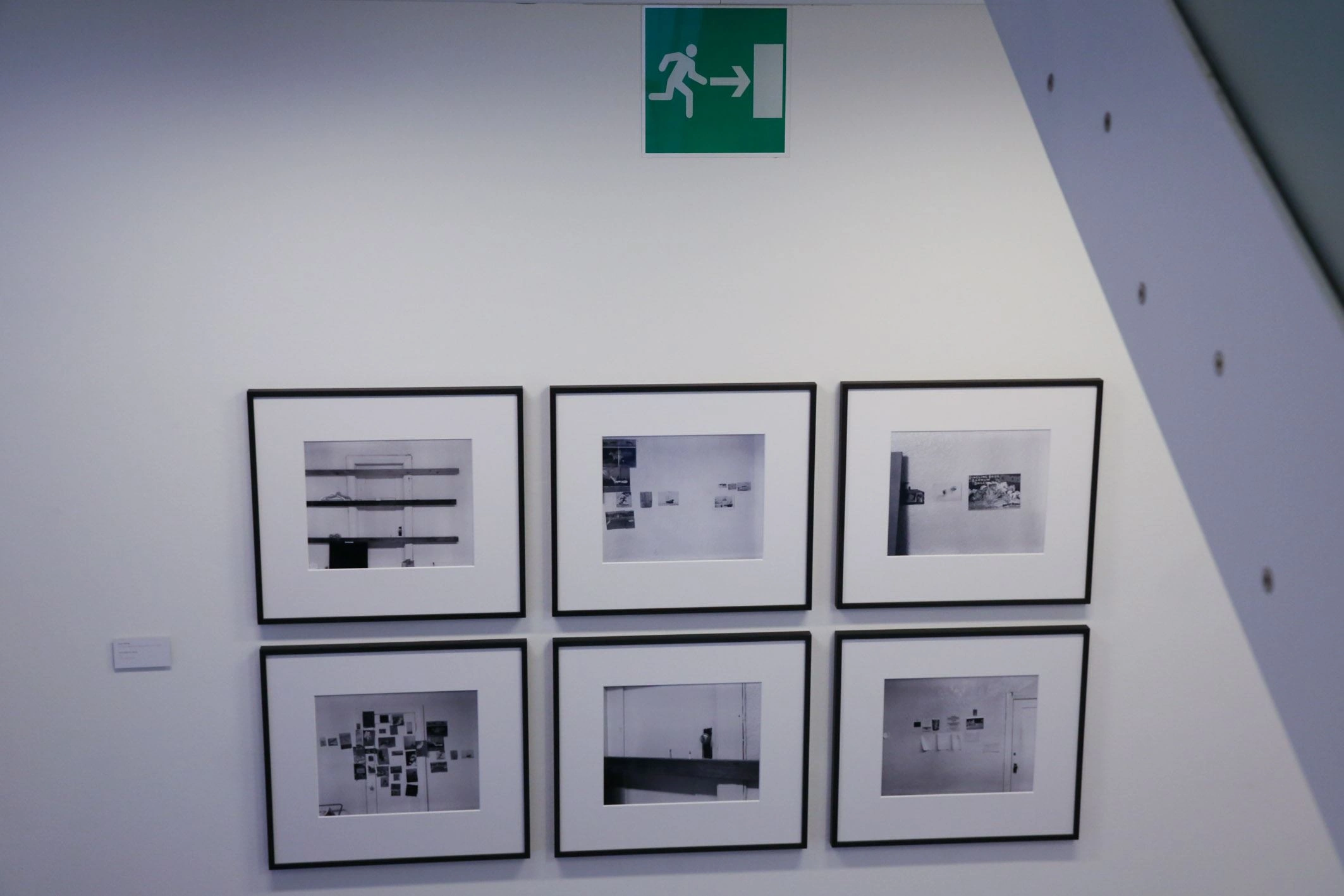 Six square pictures with passepartout frames are hanging in a stairwell. They are black and white photographs. Above them is a green sign indicating the emergency exit.