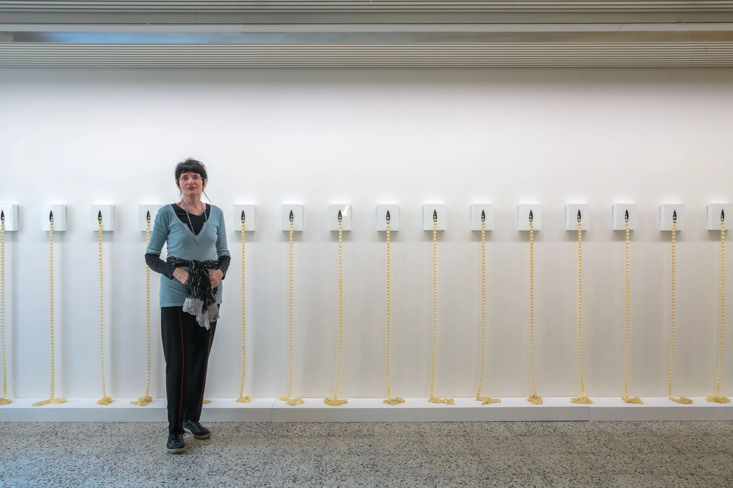 Woman standing in an art gallery next to an installation featuring a series of identical vertical elements on an illuminated wall.