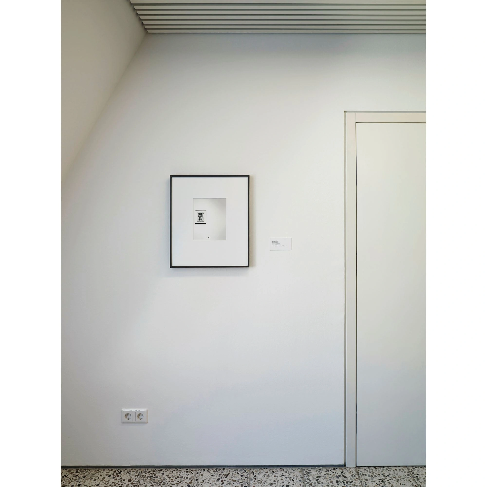 Eine Wand mit Dachschräge links ist zu sehen auf der eine Fotographie mit Passepartoutrahmen hängt. Rechts davon ist ein Türrahmen mit geschlossener Türe.