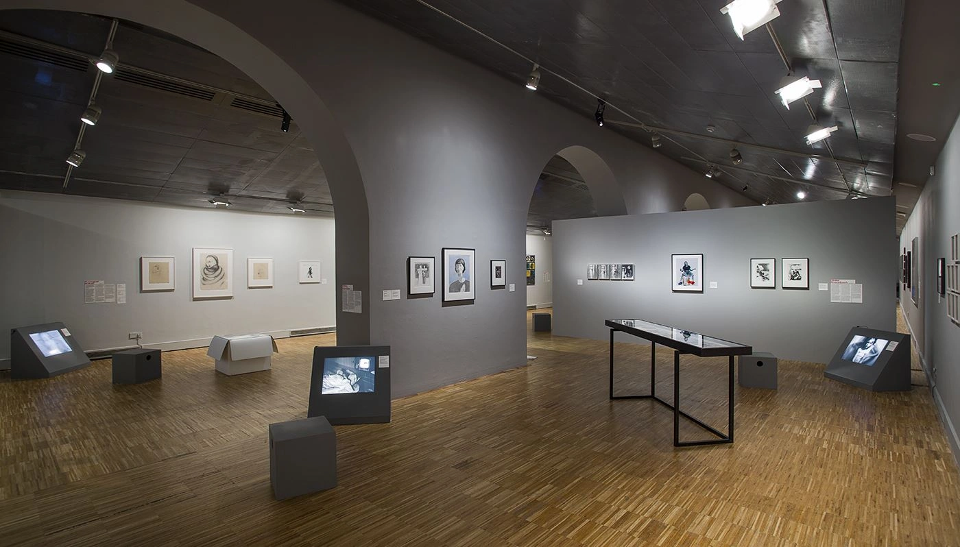 Interior view of an art gallery with vaulted ceilings and parquet floors, featuring several framed drawings on the walls and digital screens on pedestals throughout the room, under the bright illumination of ceiling spotlights.