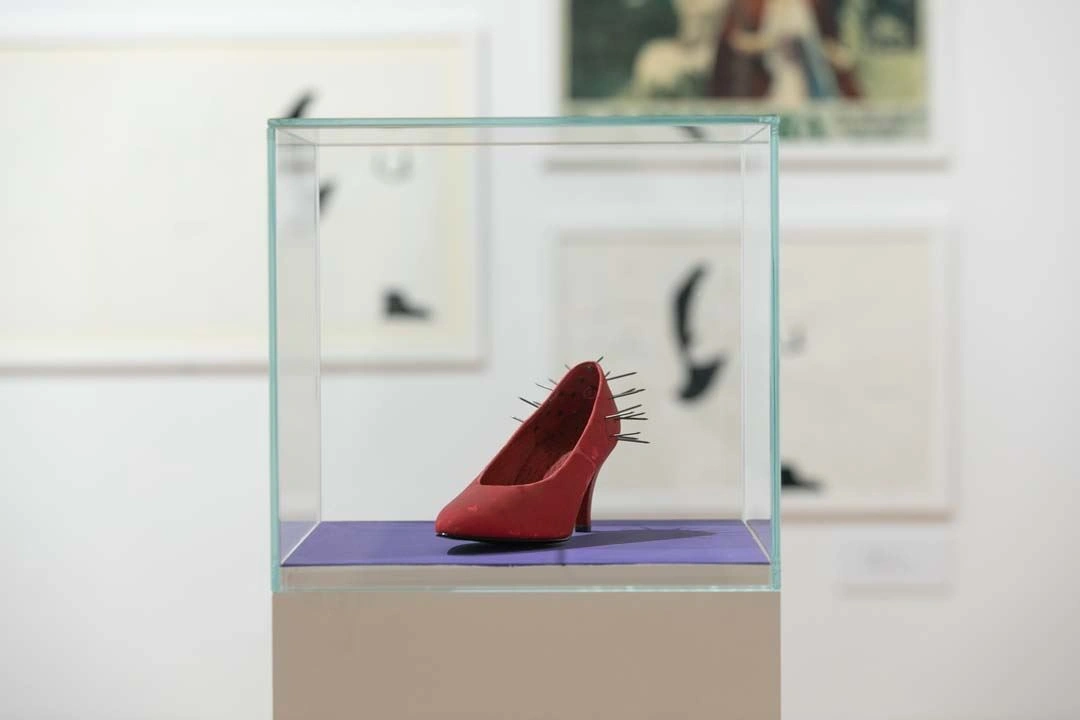 Red women's pump shoe with protruding nails on display under a glass vitrine in an art installation, with blurred black-and-white artworks in the background.