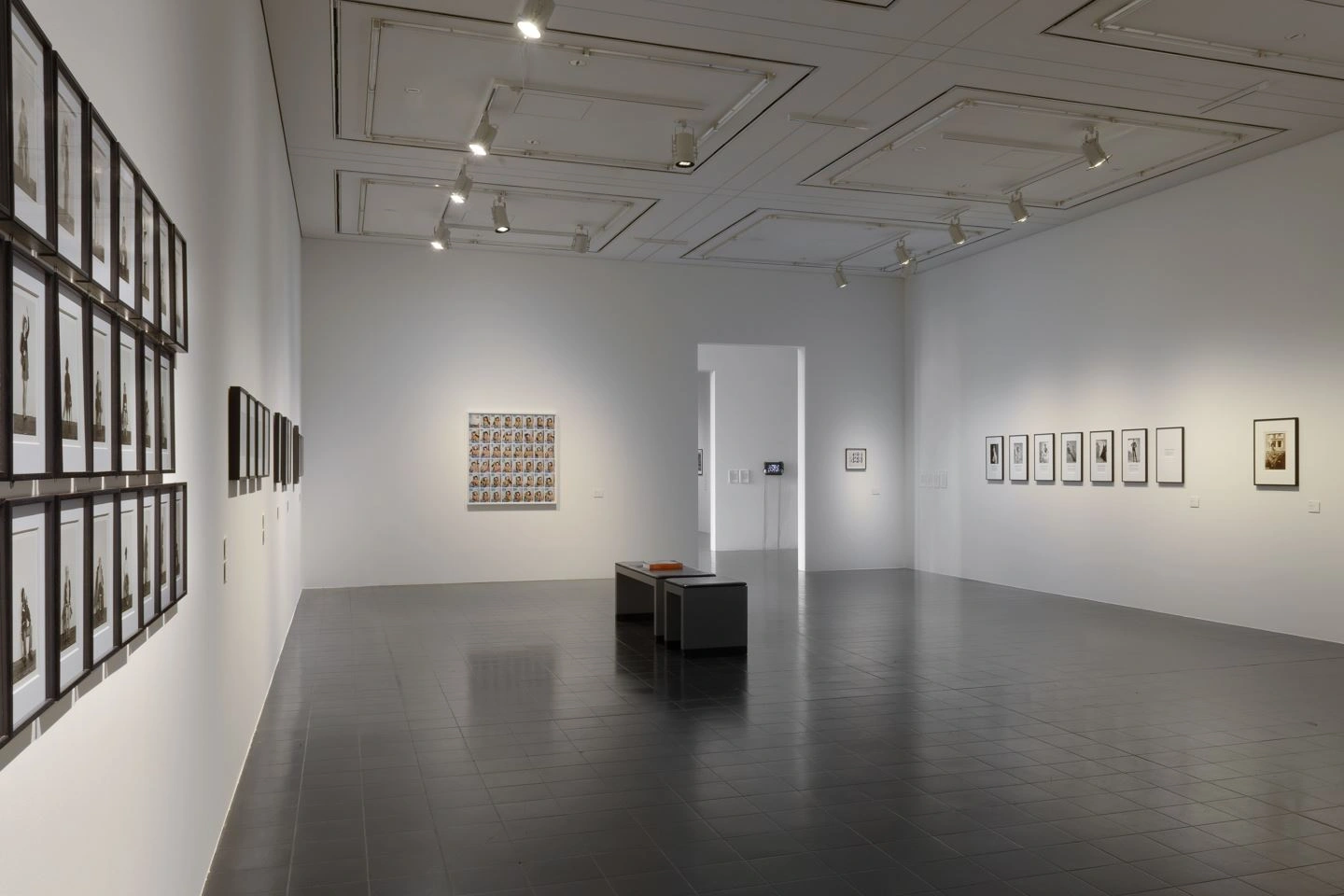 A modern exhibition room darkened can be seen. The floor looks dark grey. Framed photographs hang on all the walls. At the back right is a passageway into another room, which is brightly lit. In the middle of the room is a bench.