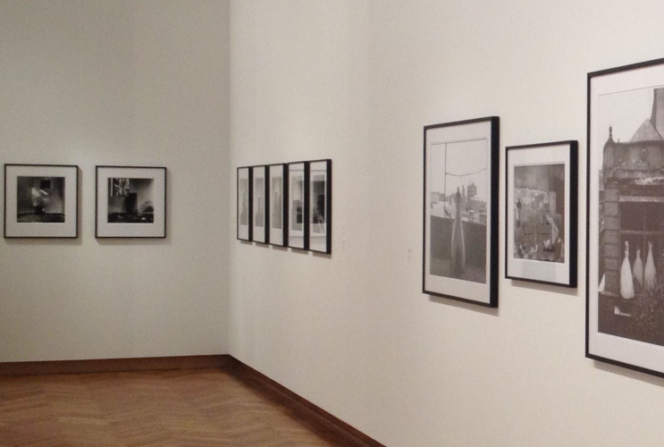 Interior view of a photo gallery featuring a selection of framed black and white photographs hung in chronological order on a white wall, in a room with wooden flooring and soft lighting.