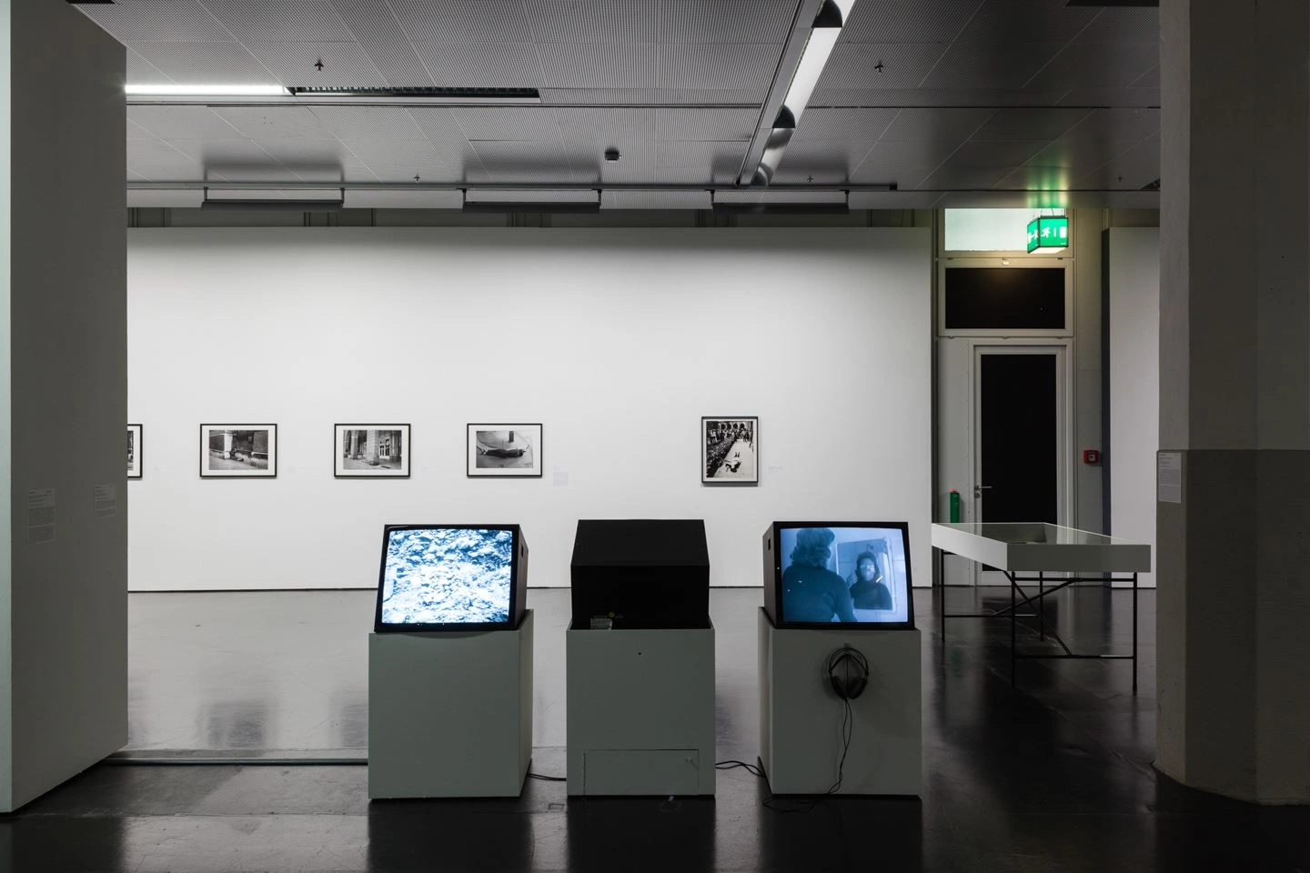 Ein Ausstellungsraum mit gedämmten Licht ist zu sehen. In der Mitte sind drei Monitore aufgestellt. Zwei davon spielen ein Video ab. Der Monitor in der Mitte ist ausgeschaltet.