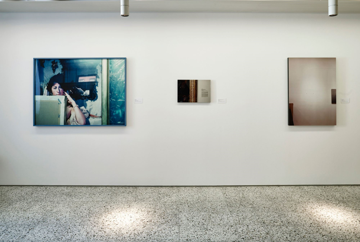 Three works of art hang on a white wall. To the left and right are large-format paintings and in the middle a small-format painting. Spotlights are placed on the ceiling that shine on the marbled floor.