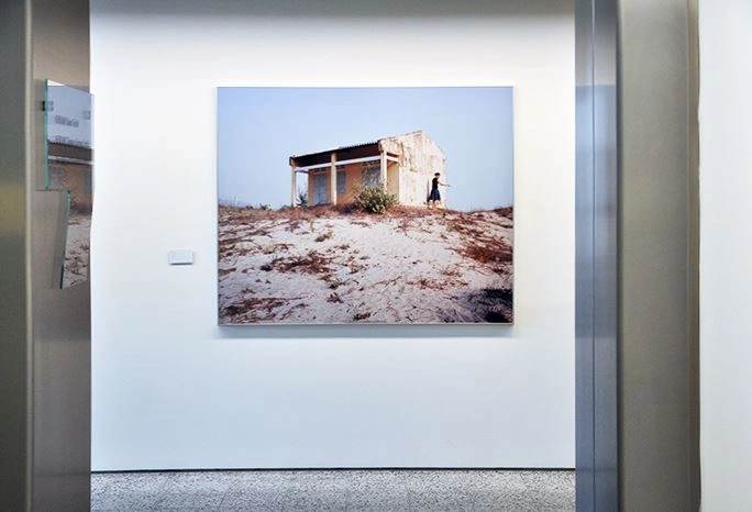 Fotografie eines verlassenen Gebäudes in einer trockenen Landschaft ausgestellt in einer modernen Kunstgalerie, flankiert von einer Glastür rechts im Bild und informativer Beschriftung links unten.