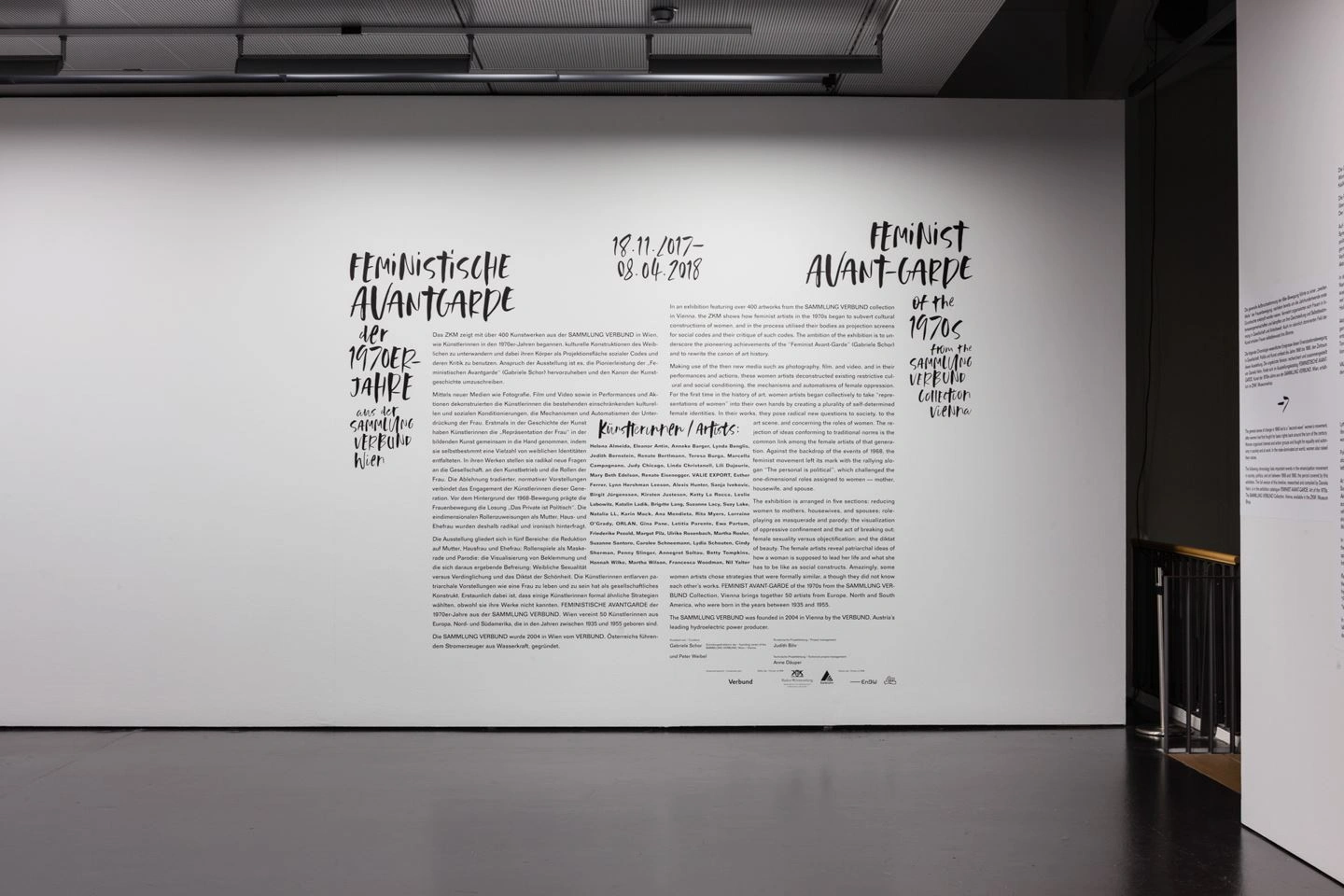 An exhibition room with a white wall and dark floor can be seen. On the white wall, a long text on the subject of feminist avant-garde can be seen in black lettering.