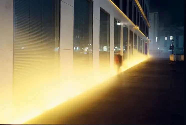 View of the VERBUND building in the courtyard. It is dark and Olafur Eliasson's installation transforms the square and the building into a stage of fog, light and wind. A shadowy figure passes through the fog.