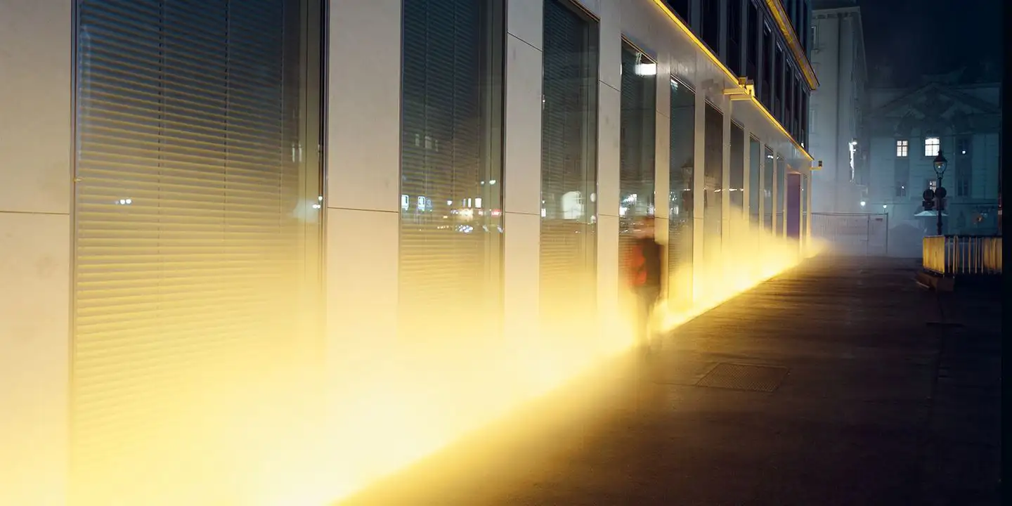 View of the VERBUND building in the courtyard. It is dark and Olafur Eliasson's installation transforms the square and the building into a stage of fog, light and wind. A shadowy figure walks through the fog.