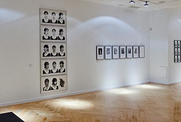 Ein moderner Ausstellungsraum im Panoramawinkel ist zu sehen. Auf der breiten Wand sind zahlreiche Kunstwerke zu sehen. An der Decke sind Lichtspots, die auf die Kunstwerke leuchten.