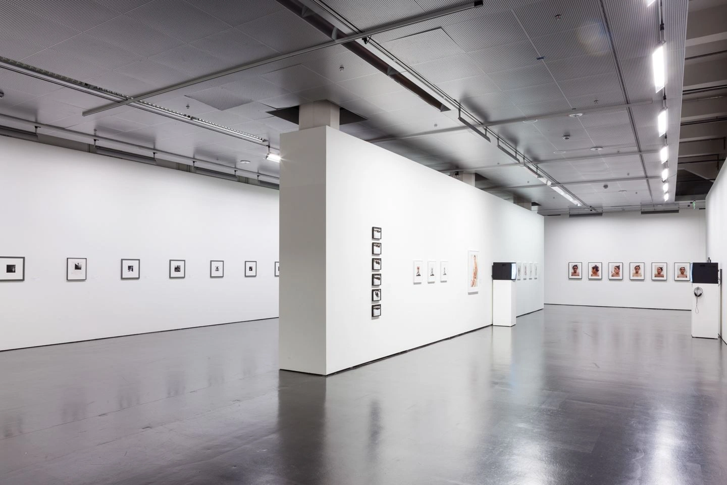 A large exhibition room can be seen. In the middle, the room is separated by a wall. Photographs hang on the walls and there are two monitors in the right part.