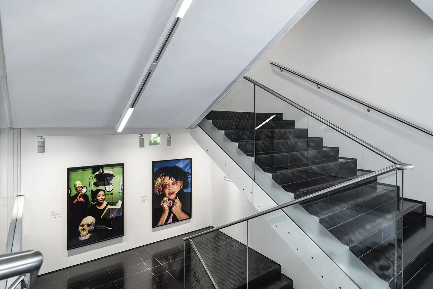 Wir blicken von oben in das Stiegenhaus der Vertikalen Galerie. In einer unteren Ebene sehen wir zwei Fotographien von ORLAN. Diese zeichnen sich durch intensive schwarze Flächen, die im Kontast zu grünen beziehungsweise blauen Flächen stehen.