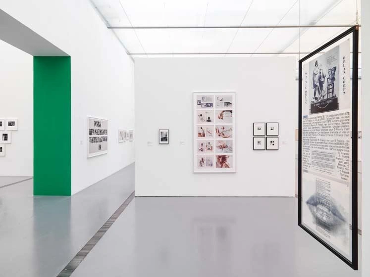 A bright exhibition room with very cold light can be seen. On the wall are photographs and other prints. The side wall of a passageway is painted in bright green.