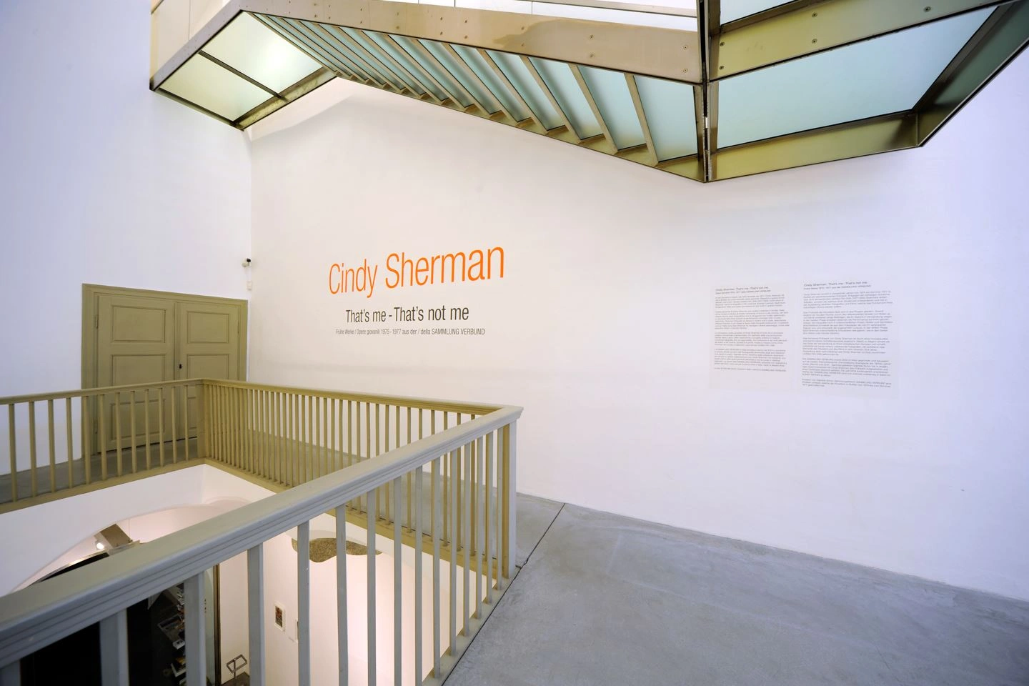 A staircase with a white wall and beige-colored stairs and wooden door is depicted. The artist's lettering and the name of the exhibition can be read on the white wall.