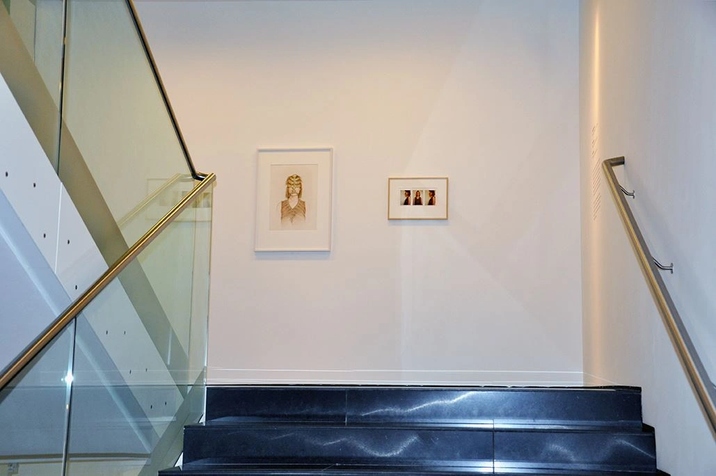 Modern black staircase with glass balustrade leading to a white wall with two framed artworks, accompanied by handrails on both sides and a concrete wall to the right.