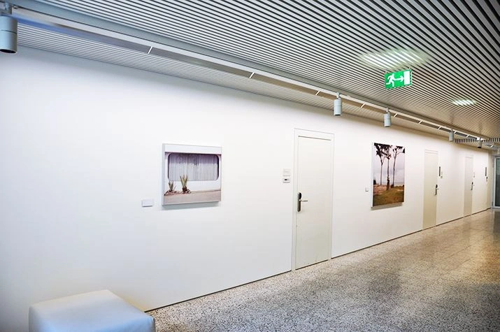 Modern gallery with a long white wall displaying two framed landscape photographs, a white bench in the foreground, and fluorescent lights on the ceiling.