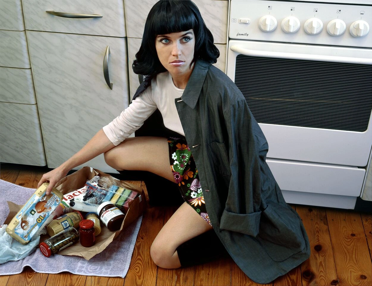 The untitled work by Aneta Grzeszykowska from 2006 shows a woman kneeling in a kitchen in front of a stove. A paper bag lies in front of her. It is torn open and the shopping is visible. She is looking at the viewer from below.