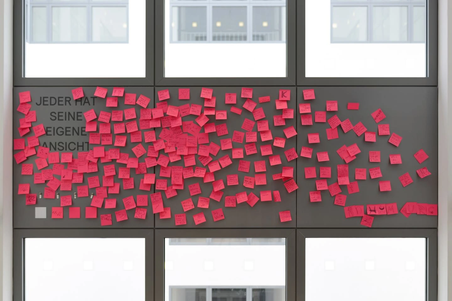 An einem grauen Fensterrahmen hängen zahlreiche rote Post-its verteilt. Dazwischen ist ein Schriftzug zu lesen: Jeder hat seine eigene Ansicht.