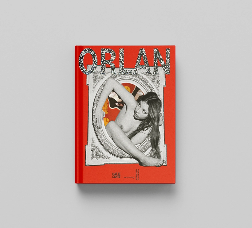 Auf einem grauen Untergrund liegt der Ausstellungskatalog der Künstlerin ORLAN. Dieser ist größtenteils rot gestaltet. Das Cover ist eine schwarz-weiß Fotografie der Künstlerin in jüngeren Jahren. Sie ist nackt und befreit sich aus einem Rahmen. Der Schriftzug ORLAN ist in glitzernden Lettern darüber angebracht.