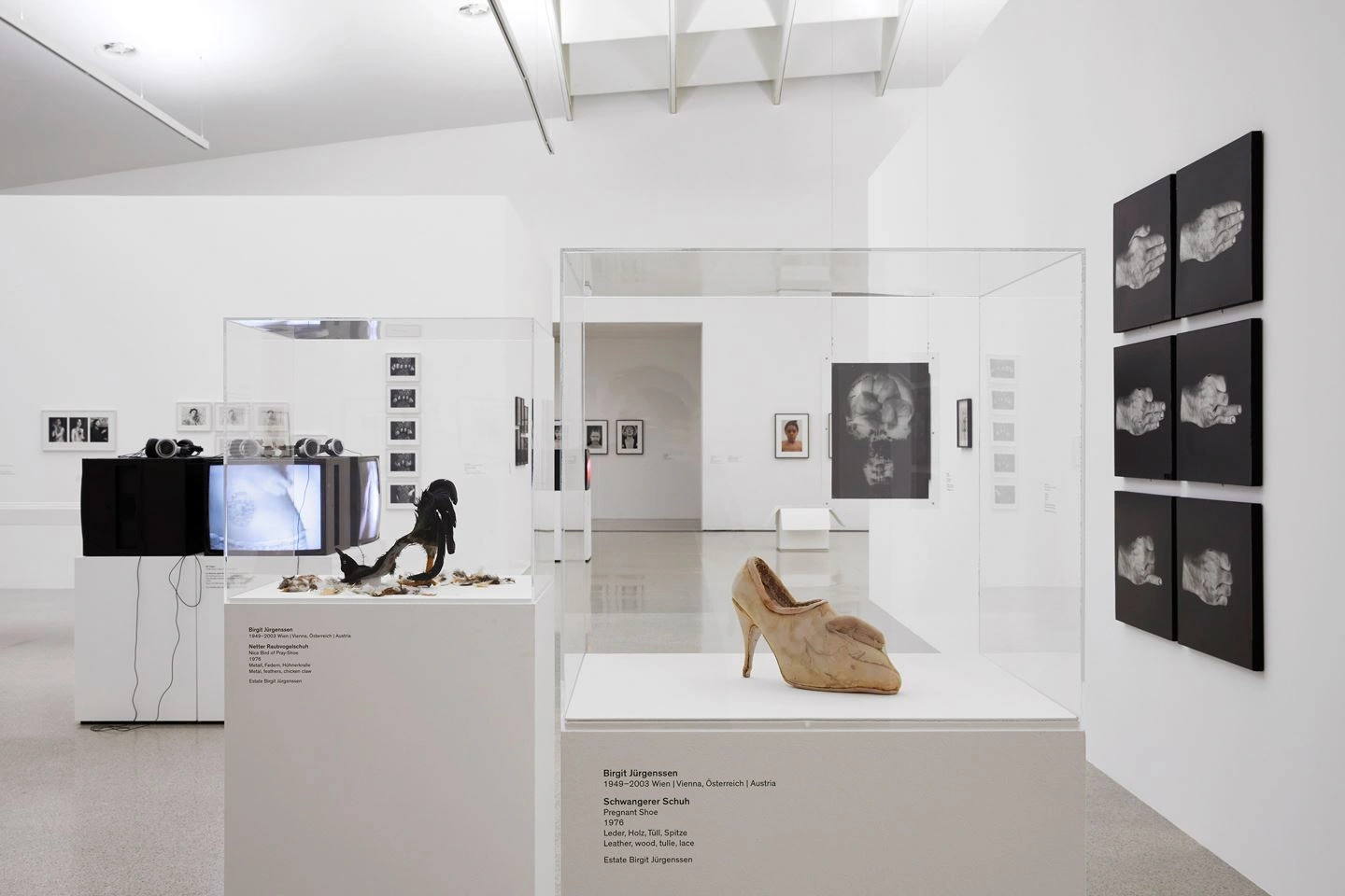 You can see an exhibition room with many exhibits. On the wall to the right are photographs on canvas with a black background and the back of a hand as a motif. On the left are monitors and in the middle are display cases with abstract objects.