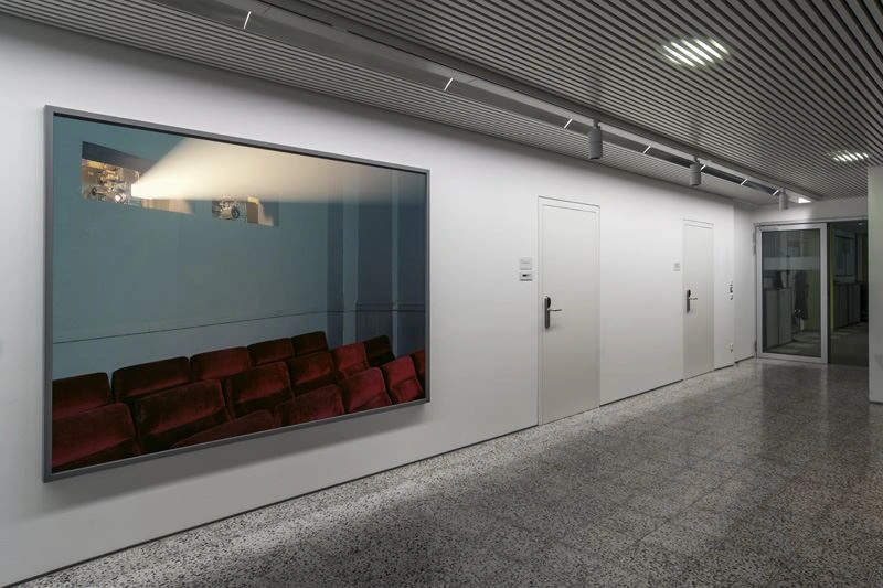 A darker office corridor with a marbled floor. A large picture in landscape format shows a movie theater with rows of seats covered in red and a projector light.