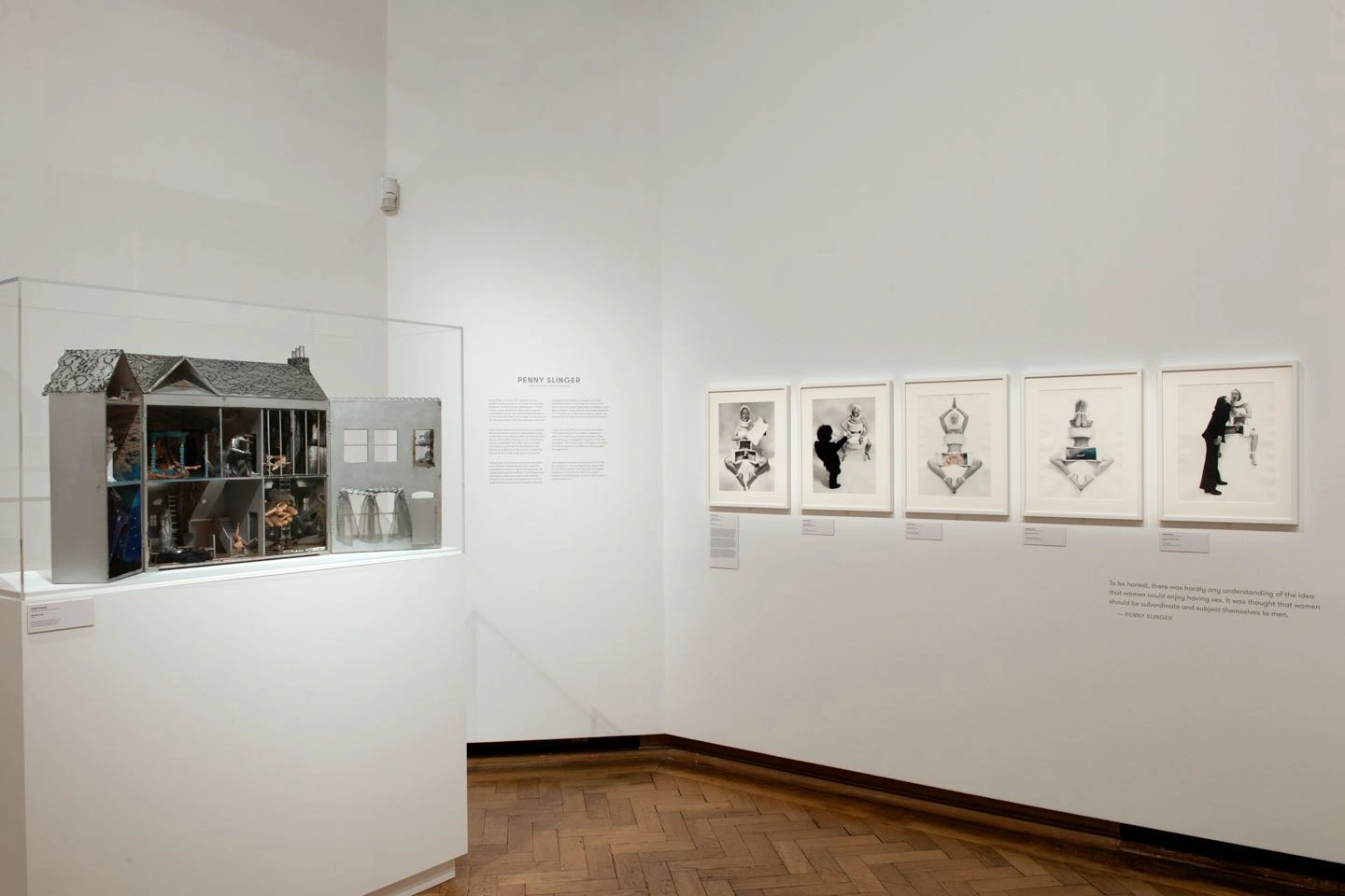 In an exhibition room of the Bozar exhibition in Brussels, there is a display case on the right with a model of a house whose interior is visible. On the right, on a white wall, there are five photographs in white picture frames.