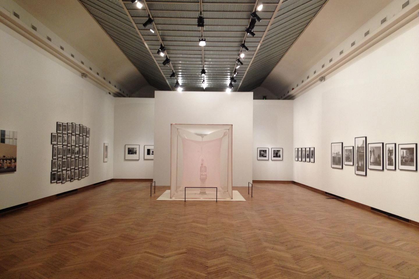 Minimalistische Ausstellungsraumgestaltung in einer Kunsthalle mit einer zentralen Vitrine, die eine Skulptur enthält, flankiert von verschiedenen gerahmten Werken an den Wänden und moderner Beleuchtung unter einer hohen Decke auf hellem Parkettboden.